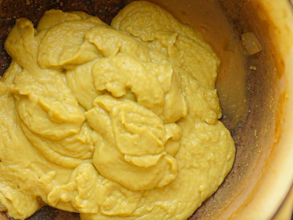 A close-up of a bowl filled with thick, smooth paste for creamy broccoli soup. The surface is uneven, showing the texture of the mustard. The bowl is partially visible, with a light brown interior contrasting against the mustard.